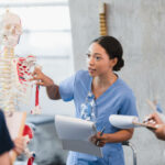 The young adult woman takes her turn to teach a class on the human skeleton as classmates take notes.