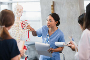 The young adult woman takes her turn to teach a class on the human skeleton as classmates take notes.